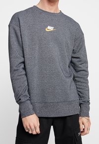 Muž v tmavě šedé mikině Nike se žlutým logem swoosh, černých kalhotách a viditelnými tetováními na rukou, na jednobarevném pozadí.