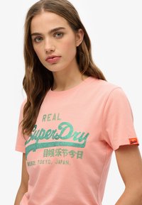 T-shirt rose à manches courtes avec un logo "SuperDry" vert pailleté et un texte japonais, accompagné d'une petite étiquette orange sur la manche.