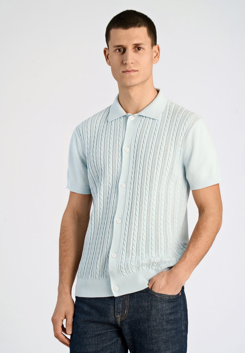 Vaaleansininen lyhythihainen neulejakku, jossa on kaapelineulekuvio, etupainike ja ribbitetyöt kauluksessa ja helmassa.