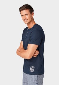 Sötétkék pamut póló rövid ujjakkal és kerek nyakkivágással, három gombos kivágással és fehér logó részlettel az alján.