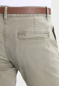 Pantalons kaki en tissu léger, avec une ceinture en cuir marron, une poche arrière avec un rabat et une texture subtile.