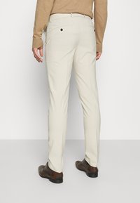 Bézs, slim fit szabású nadrág, sima anyagból készült. Két hátsó zsebe van fekete gombokkal és klasszikus derékrésszel.