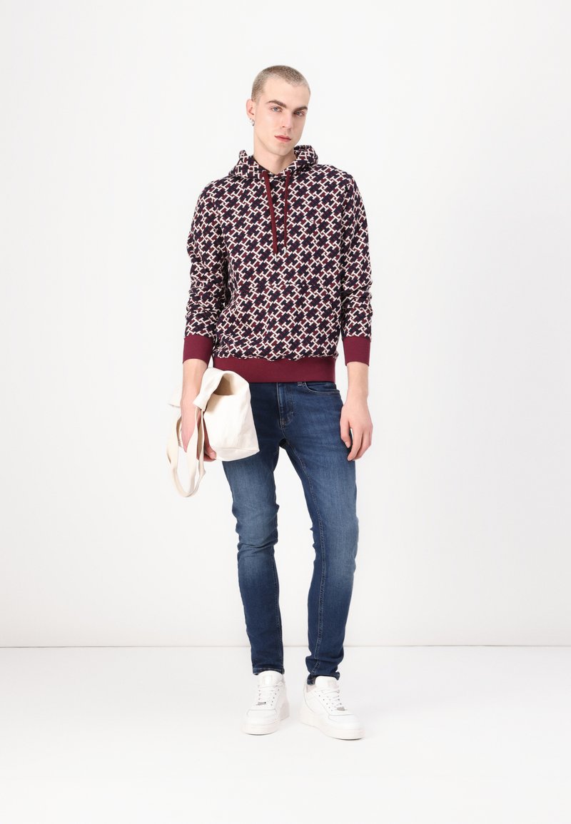 Felpa a giacca con cappuccio a disegni marrone e bianco, abbinata a jeans blu e sneakers bianche. Tiene una borsa color crema.