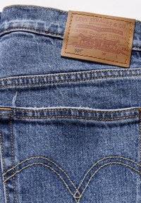 Zadní kapsa modrých džín Levi's 501 s podpisovým koženým štítkem a detaily oranžového prošívání na denimové tkanině.