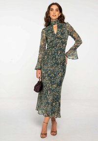 Mujer con un vestido largo, estampado en verde y marrón, con mangas acampanadas, sosteniendo un pequeño bolso oscuro y usando zapatos de tacón dorados con tiras.