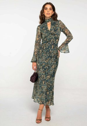 Mujer con un vestido largo, estampado en verde y marrón, con mangas acampanadas, sosteniendo un pequeño bolso oscuro y usando zapatos de tacón dorados con tiras.