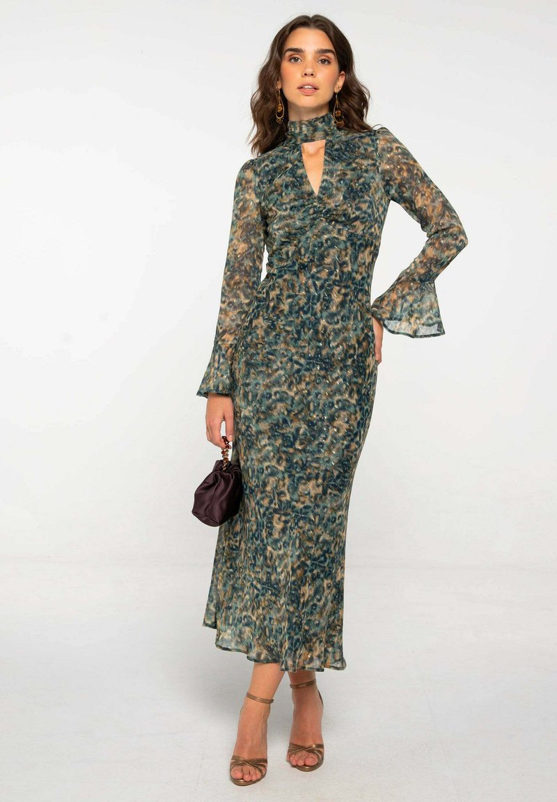 Mujer con un vestido largo, estampado en verde y marrón, con mangas acampanadas, sosteniendo un pequeño bolso oscuro y usando zapatos de tacón dorados con tiras.