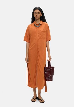 Kobieta o długich czarnych włosach, ubrana w pomarańczową midi sukienkę zapinaną na guziki, czarne sandały, masywny naszyjnik i trzymająca bordową skórzaną torebkę.