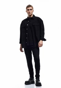 Camisa chaqueta oversized negra con dos bolsillos frontales, llevada sobre una camiseta negra y combinada con pantalones negros ajustados y botas negras gruesas.