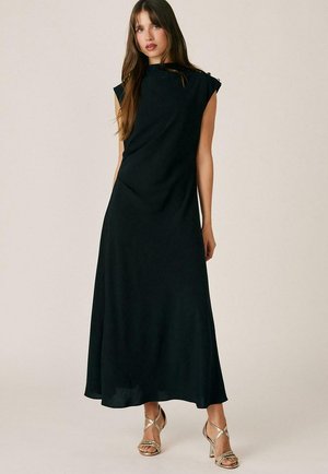 Joven mujer con el pelo largo, vestida con un vestido maxi negro sin mangas y sandalias doradas de tacón alto con tiras, de pie sobre un fondo liso.