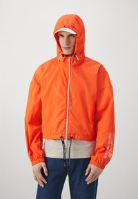 Orange cropped jakke med hætte, frontlynlås, elastiske manchetter og snoretræk i kanten. Har hvid logo tryk på ærmet og en glat tekstur.