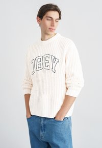 Pulover tricotat de culoare crem cu textură striată, având inscripția "OBEY" în litere negre pe partea din față. Purtat cu blugi albaștri din denim.