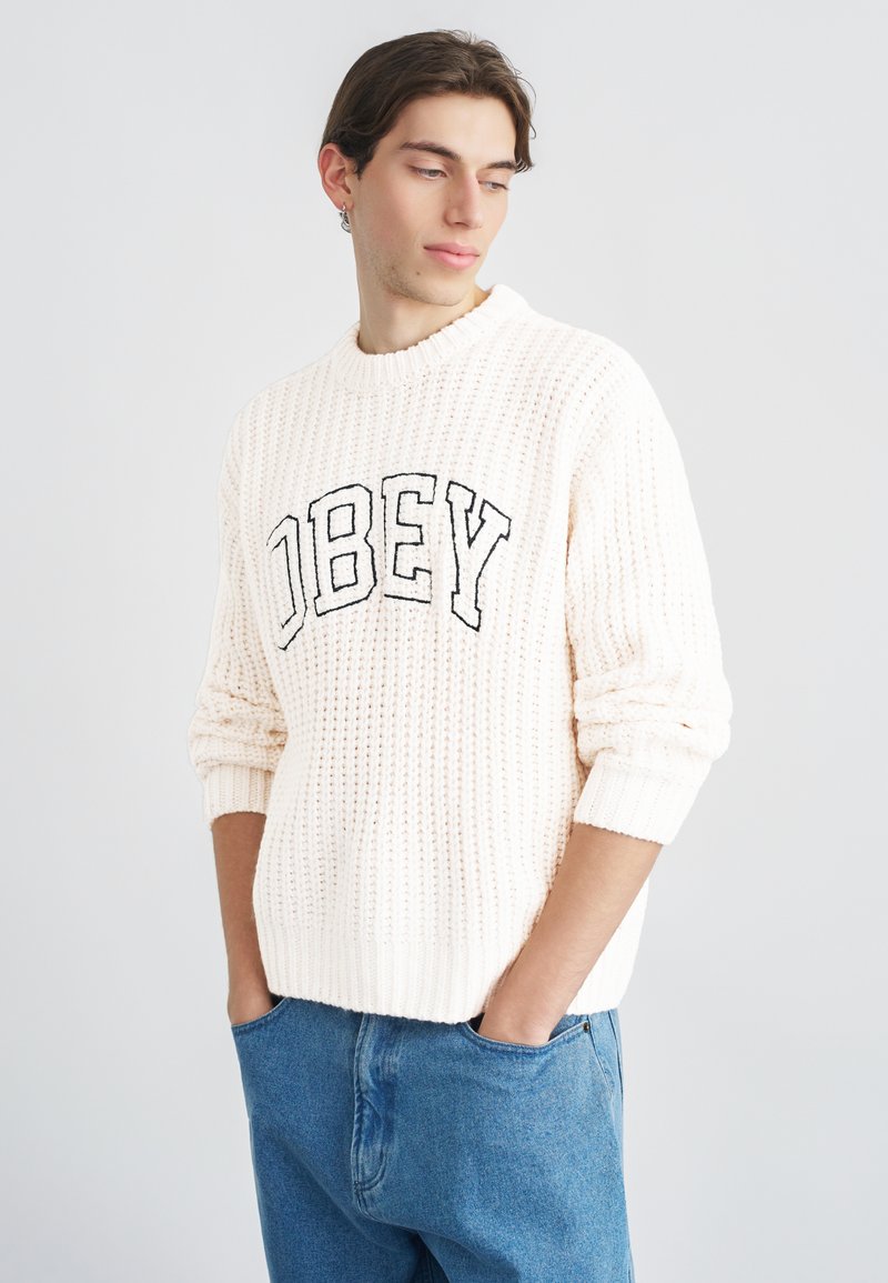 Pulover tricotat de culoare crem cu textură striată, având inscripția "OBEY" în litere negre pe partea din față. Purtat cu blugi albaștri din denim.