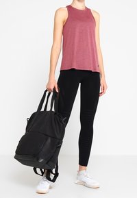Violetti toppi, mustat legginssit, joissa on tekstuurisia yksityiskohtia, ja musta reppu etutaskulla, jonka kantaa henkilö valkoisissa lenkkareissa.