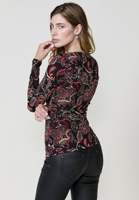 Dug majica s dugim rukavima s crnom osnovom i crvenim i zlatnim cvjetnim paisley uzorkom. Prilagođen dizajn s okruglim izrezom i manžetama na zapešćima.