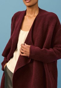 Burgunderfarbener, übergroßer Cardigan aus strukturiertem Stoff, mit offenem Design und drapiertem Vorderteil. Wird über einem cremefarbenen Oberteil getragen, vor einem blauen Hintergrund.
