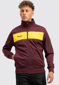 Bordó zipzáras dzseki sárga csíkkal a mellkason, magas gallérral, bordázott mandzsettákkal és a "Lonsdale" logóval.