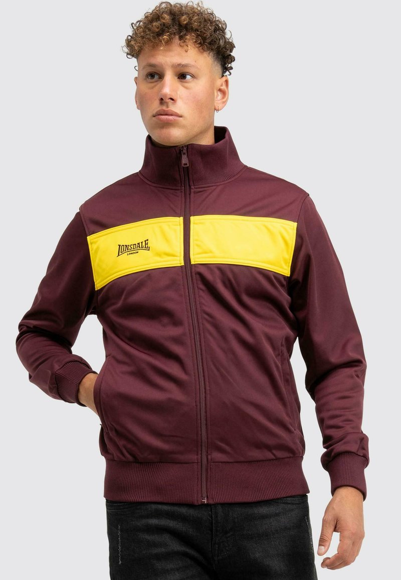 Bordó zipzáras dzseki sárga csíkkal a mellkason, magas gallérral, bordázott mandzsettákkal és a "Lonsdale" logóval.