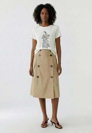 Femme aux cheveux bouclés portant un t-shirt blanc à motif, une jupe midi beige boutonnée et des sandales à talons marron, debout devant un fond uni.