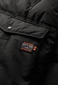 Sort isoleret jakke med en stor frontlomme, trykknap og et orange og gråt logo-patch mærket "Superdry Expedition."