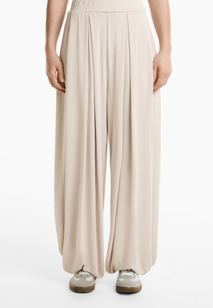 Kerman beige leveled wide-leg pants dengan waistband tinggi, tekstur halus, dan dipadukan dengan sepatu sneakers abu-abu dan putih.