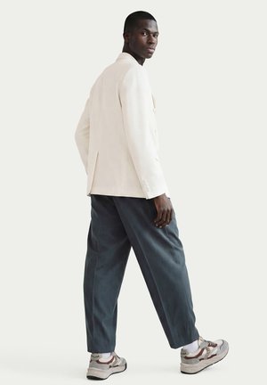 Uomo con blazer beige, pantaloni larghi grigio scuro e sneakers bianche, che guarda oltre la spalla su uno sfondo chiaro e uniforme.