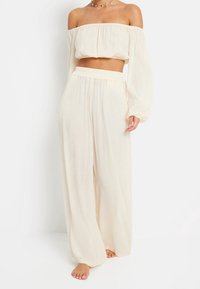 Woman wearing cream off-shoulder long-sleeve crop top and matching high-waisted wide-leg pants, barefoot, against white background.