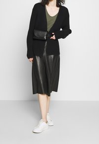 Svart virkad cardigan över en olivgrön topp, kombinerad med en svart läder midi-kjol och vita sneakers. Outfiten har en svart axelremsväska.