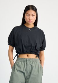 Black cropped t-shirt with short, puffed sleeves, drawstring hem, and a small logo detail at the neckline. Paired with green cargo pants.