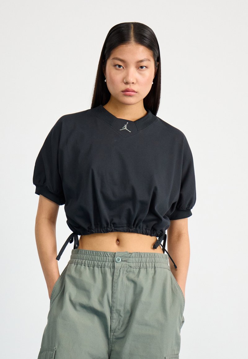 Black cropped t-shirt with short, puffed sleeves, drawstring hem, and a small logo detail at the neckline. Paired with green cargo pants.