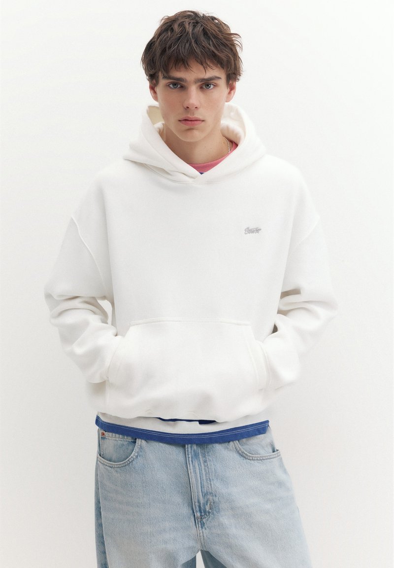 Jeune homme portant un sweat à capuche blanc, les mains dans les poches et un jean bleu clair, debout devant un fond blanc uni.