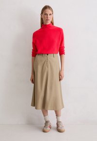 Helles rotes, hochgeschlossenes Sweatshirt kombiniert mit einem khakifarbenen, knielangen Rock. Abgerundet mit beigen Sneakers und weißen, gerippten Crew-Socken.