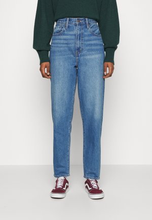 Jeans taille haute bleu clair avec une coupe droite, présentant une légère décoloration et cinq poches, associés à des baskets bordeaux.
