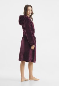 Robe con cappuccio color bordeaux in morbido tessuto plush, con vita regolabile, tasche profonde e design a lunghezza ginocchio. Presenta una texture liscia e cuciture pulite.