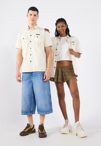 Camicia beige a maniche corte con due tasche frontali, abbinata a pantaloni corti blu a gamba larga. Modella femminile con top corto bianco e gonna a quadri.