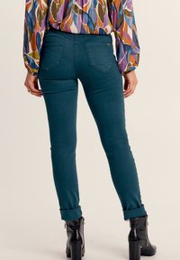 Jeans skinny color teal con una texture liscia e passanti per la cintura, dotati di due tasche posteriori, con orli arrotolati e abbinati a stivali neri con tacco.