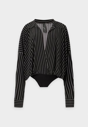 Femme aux cheveux bouclés porte une veste matelassée noire brillante, une chemise rayée à col en V profond, un pantalon noir à jambes larges et des chaussures noires pointues.