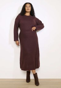 Burgundijas adīta kleita ar garām piedurknēm, pieguļošu jostasvietu un taisnu svārku. Saskaņota ar tumši brūniem potīšu zābakiem.