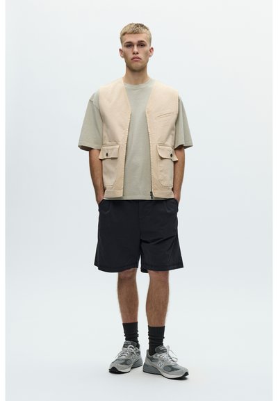 Młody mężczyzna stojący, ubrany w beżową kamizelkę, jasnoszarą oversize koszulkę, czarne szorty, czarne skarpety i szare trampki na białym tle.