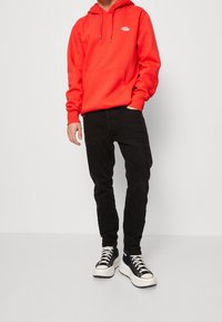 Röd hoodie med en central ficka, svarta tight jeans och svartvita sneakers. Hoodien har en logotyp på bröstet.