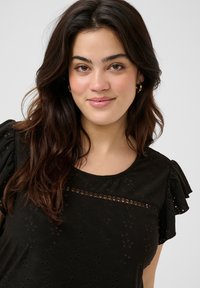 Blusa negra con mangas cortas y volantes, que presenta una tela texturizada con detalles de ojal y un panel horizontal transparente en el pecho.