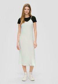 Robe slip en satin vert clair avec des bretelles blanches, superposée à une chemise noire à manches courtes, associée à des baskets blanches à semelles épaisses.