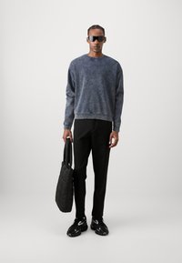 Mørkeblå hettegenser med gradienttekstur, svarte bukser og svart quiltet totebag; svarte solbriller og sneakers fullfører stilen.