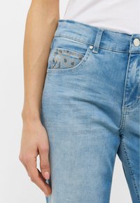 Jean en denim bleu clair à taille haute, avec un design de poche à motifs et une fermeture à bouton à l'avant. Texture lisse, style décontracté.