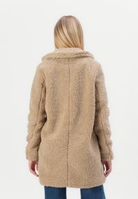 Béžový, načechraný teddy kabát s límcem, dlouhými rukávy a přímým střihem. Má texturovaný povrch a dvě boční kapsy.