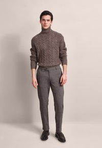 Brauner, kabelgestrickter Rollkragenpullover kombiniert mit grauen, maßgeschneiderten Hosen und schwarzen Dressschuhen. Einfaches Design und strukturiertes Material.