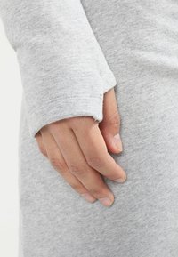 Main reposant contre une manche en tissu gris avec un trou pour le pouce, montrant une posture décontractée et une texture douce du vêtement.
