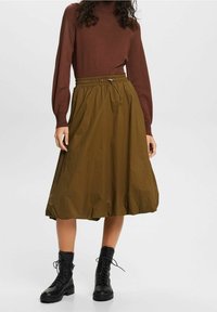 Haut marron à manches longues associé à une jupe mi-longue couleur vert olive avec une taille élastique et un cordon de serrage. L'ensemble est complété par des bottes noires à lacets.