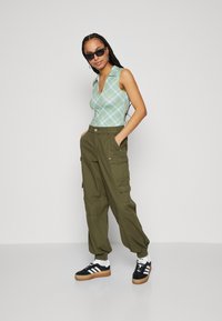 Light green plaid sleeveless top, olive cargo pants with side pockets, and black sneakers with gum soles. Sunglasses complete the look.