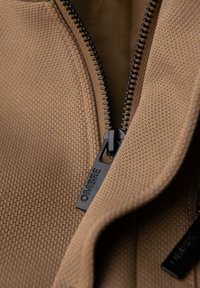 Braun strukturiertes Material mit einem schwarzen Reißverschluss, der in der Mitte verläuft. Der Reißverschluss-Zipper ist mit "OMBRA" graviert und zeigt ein einfaches Design.
